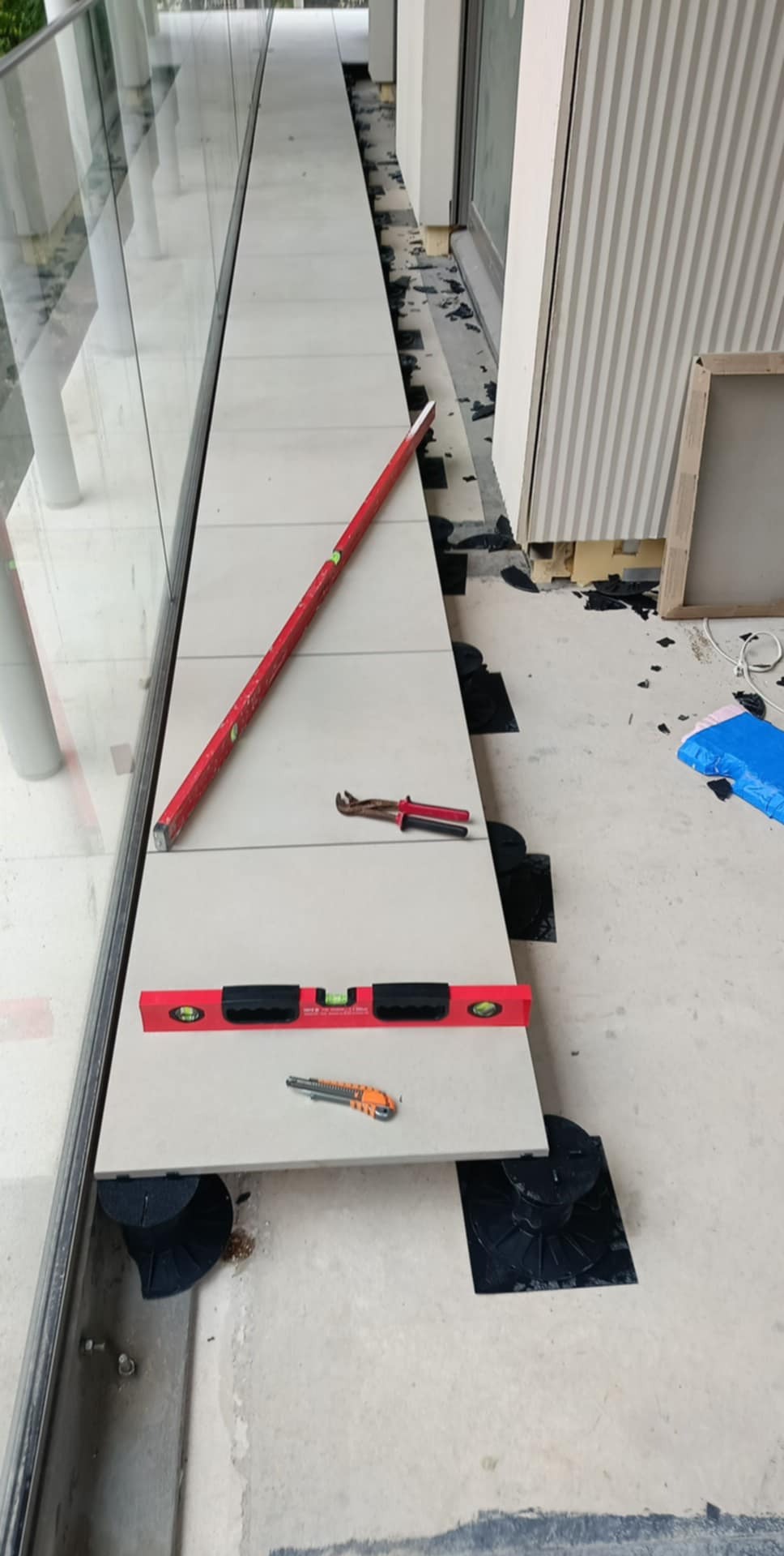 Montaż podłogi na balkonie z użyciem poziomicy i specjalnych podkładek. Widoczne narzędzia: nóż i szczypce. Prace wykończeniowe w toku, jasne płytki.