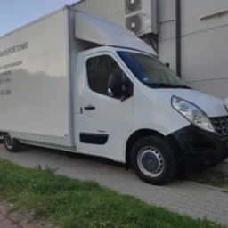 Biały bus dostawczy z napisem 'Usługi Transportowe' i numerem telefonu, zaparkowany na chodniku z kostki brukowej. Widoczne budynki w tle i trawa przy chodniku.