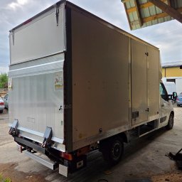 Biały samochód dostawczy z zabudową kontenerową, zaparkowany na zewnątrz, widok z tyłu i boku, z widocznymi elementami konstrukcyjnymi i tabliczką informacyjną.