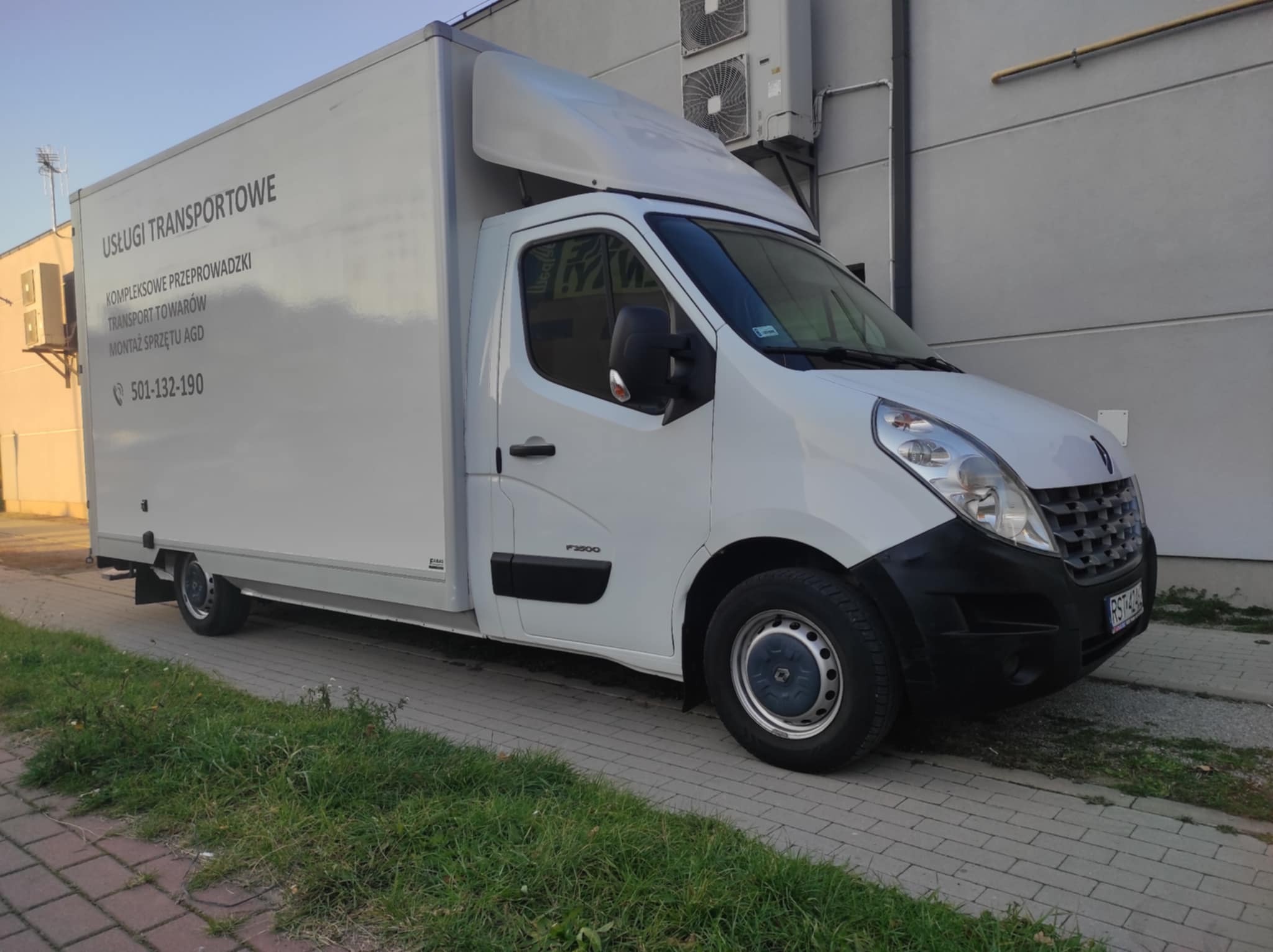 Biały bus dostawczy z napisem 'Usługi Transportowe' i numerem telefonu, zaparkowany na chodniku z kostki brukowej. Widoczne budynki w tle i trawa przy chodniku.