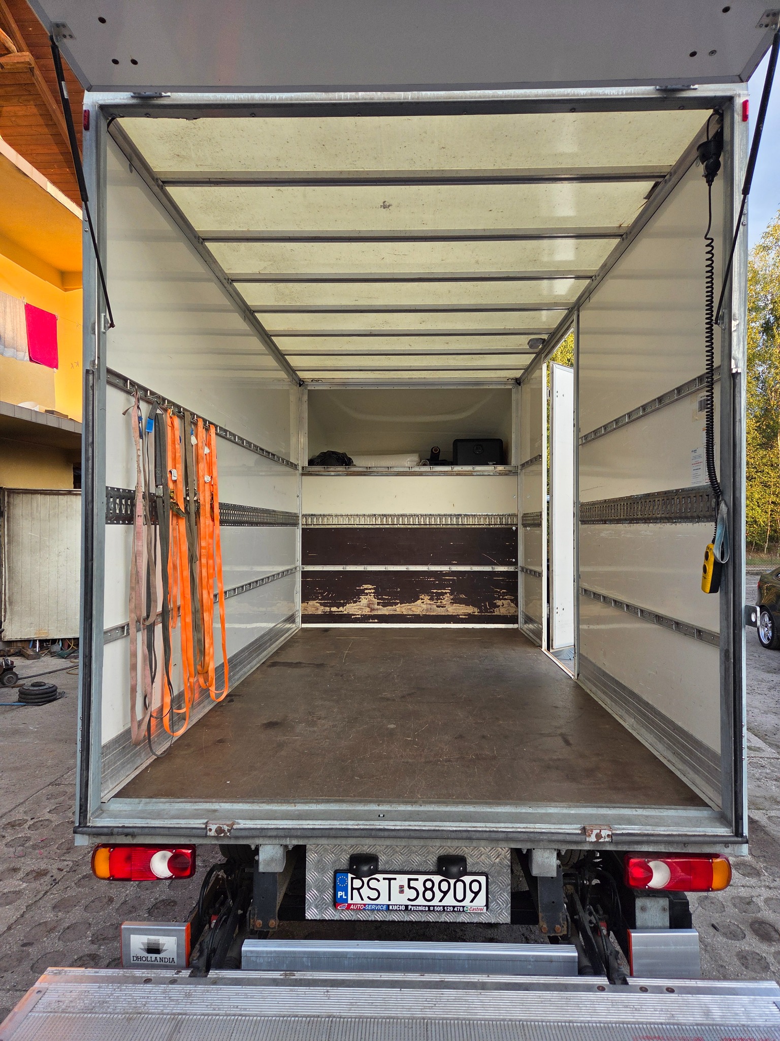 Pusta przestrzeń ładunkowa busa z pasami mocującymi, widok z tyłu, otwarte drzwi. Wnętrze wyłożone panelami, podłoga z płyty. Tablica rejestracyjna widoczna.