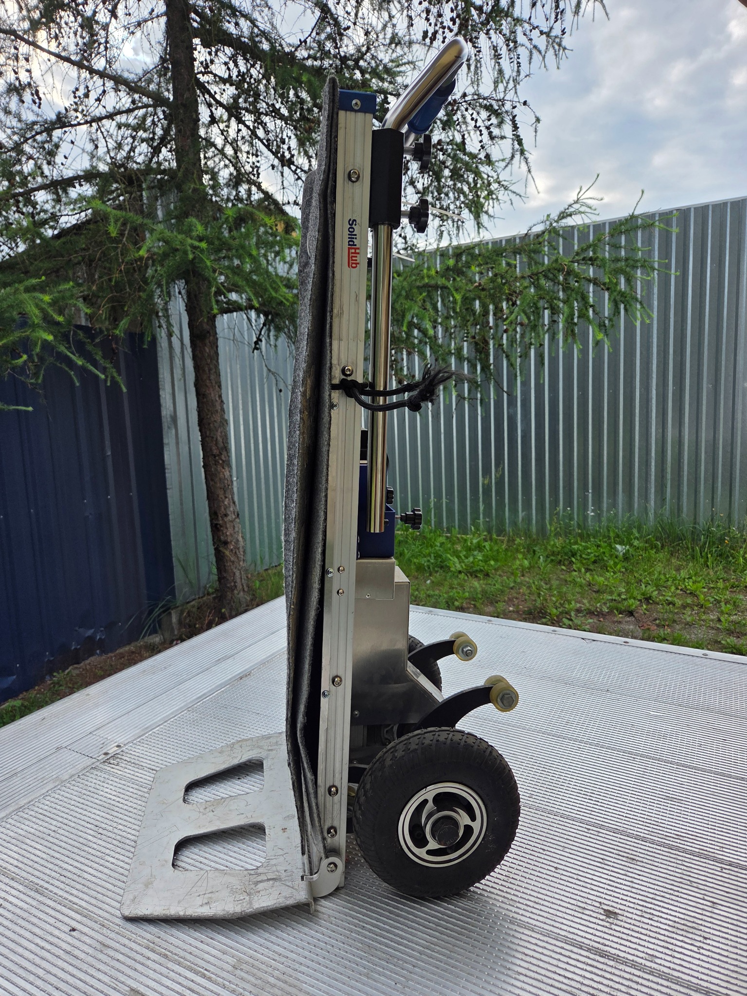 Elektryczny wózek schodowy SolidHub na platformie transportowej, z widocznym metalowym ogrodzeniem w tle i drzewem iglastym.