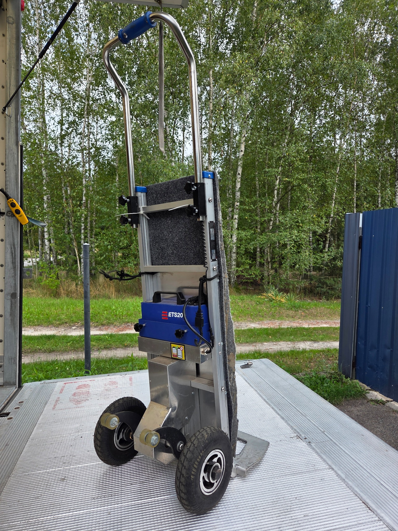 Wózek schodowy elektryczny ETS20 na platformie załadunkowej, z widocznym lasem w tle. Urządzenie gotowe do transportu towarów po schodach.