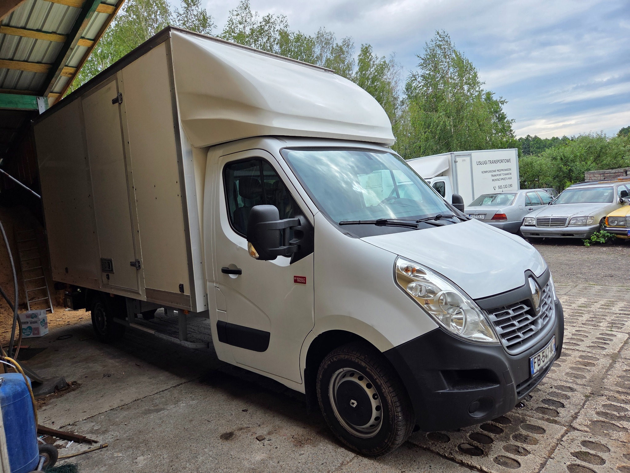 Biały bus dostawczy Renault Master z zabudową kontenerową, zaparkowany na betonowym placu. W tle zieleń drzew i inne pojazdy. Widoczny numer rejestracyjny.