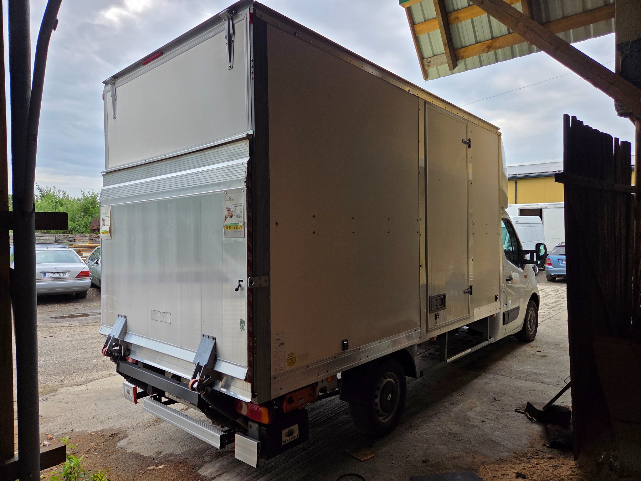 Biały samochód dostawczy z zabudową kontenerową, zaparkowany na zewnątrz, widok z tyłu i boku, z widocznymi elementami konstrukcyjnymi i tabliczką informacyjną.