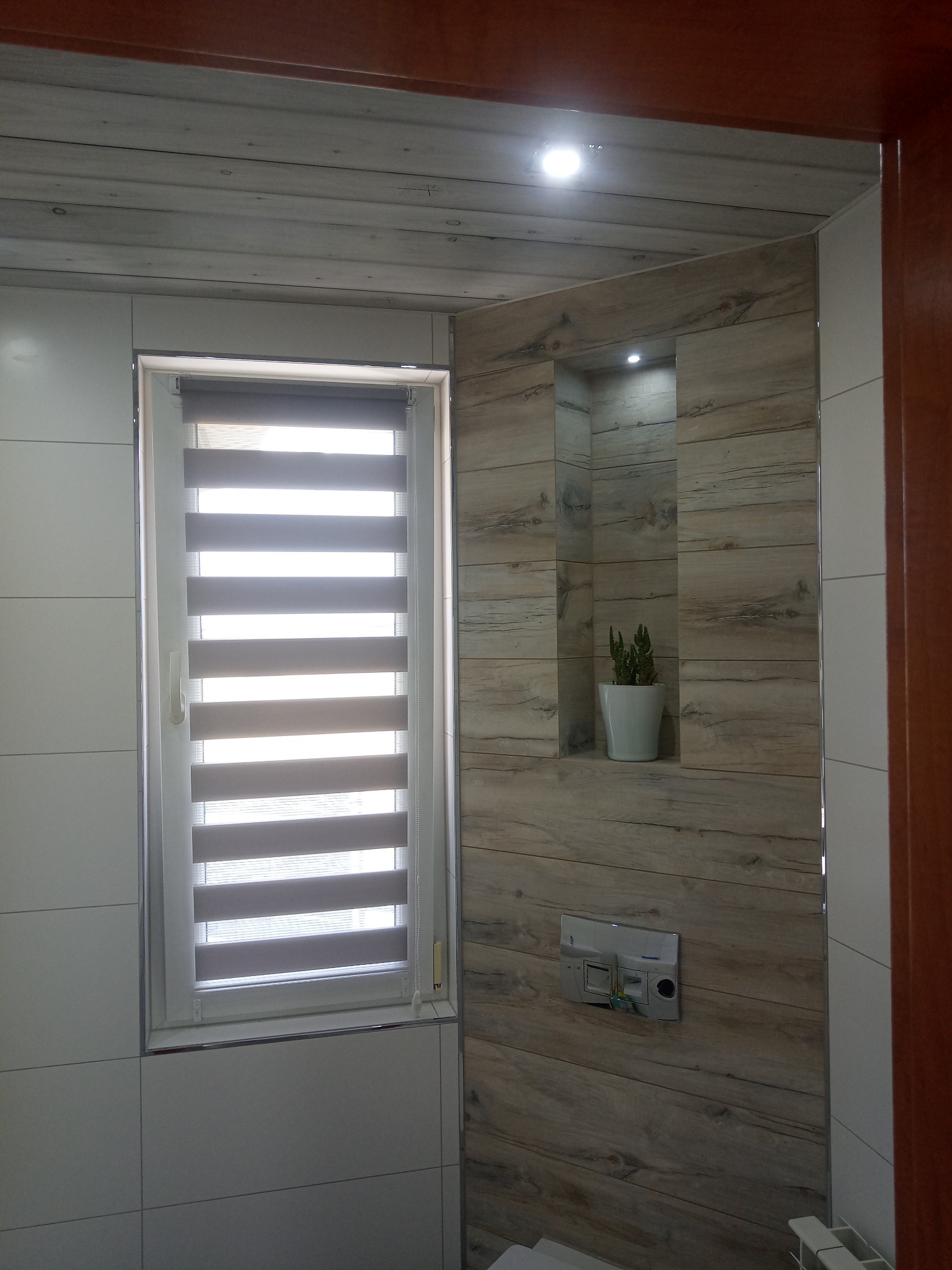 Wykończona łazienka z oknem i roletami dzień-noc. Ściany wyłożone płytkami imitującymi drewno i białymi, gładkimi. Wnęka z oświetleniem i rośliną doniczkową.