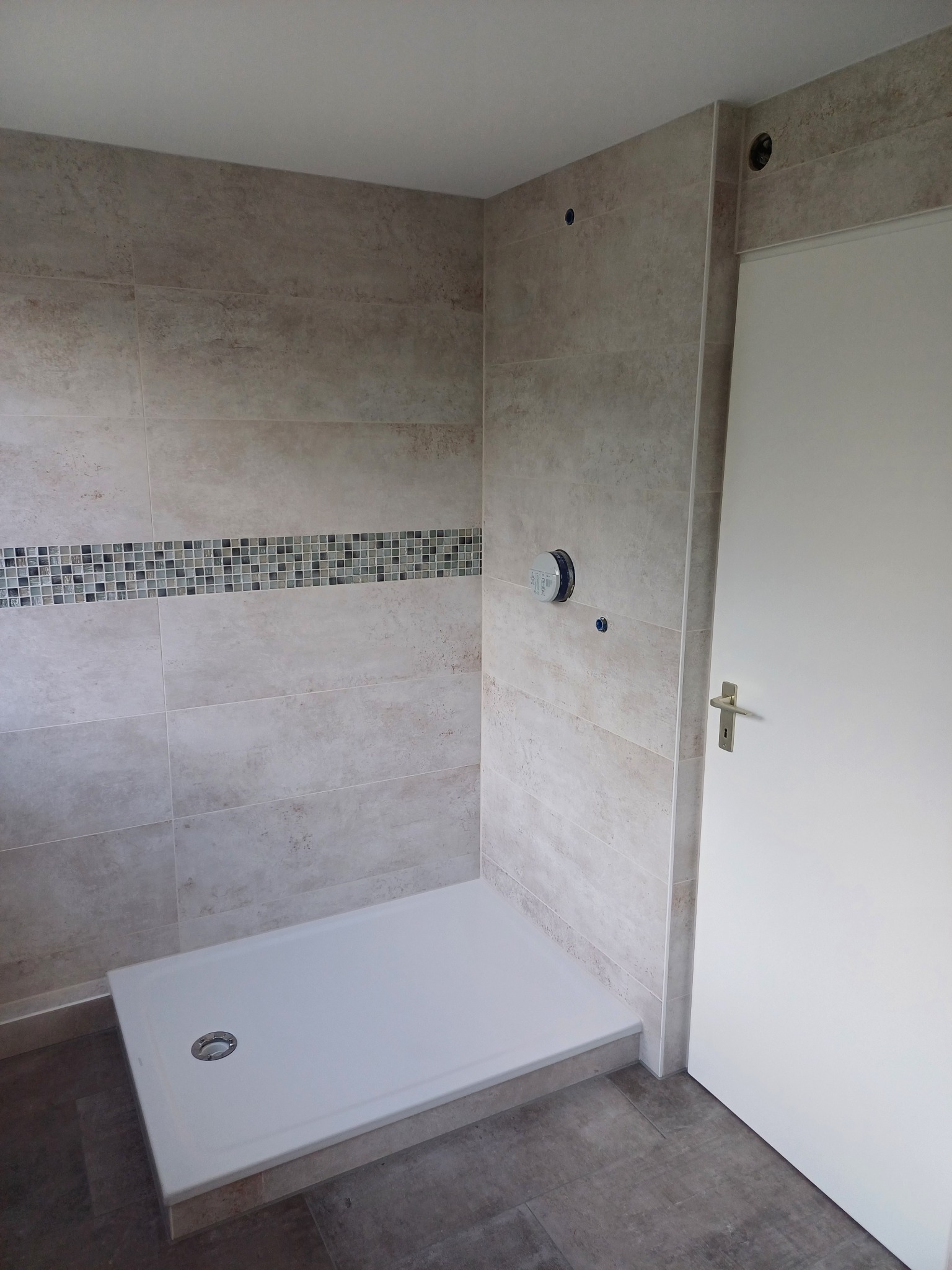 Wykończona kabina prysznicowa z beżowymi płytkami i dekoracyjnym pasem mozaiki. Biały brodzik i drzwi. Widoczne elementy instalacji.