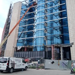 MAL-BAU WOŹNICA, KOWALCZYK SPÓŁKA CYWILNA - Prace na wysokościach przy szklanej elewacji biurowca. Podnośnik koszowy i rusztowania. Na pierwszym planie van z reklamą usług: malowanie dachów, elewacji i konstrukcji stalowych.