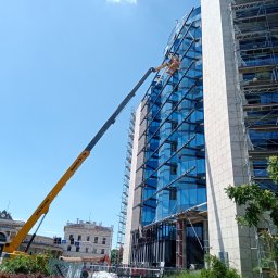 MAL-BAU WOŹNICA, KOWALCZYK SPÓŁKA CYWILNA - Praca na wysokościach: Żółty podnośnik koszowy przy szklanej elewacji biurowca w słoneczny dzień. Widoczne rusztowania i budynek dworca w tle.