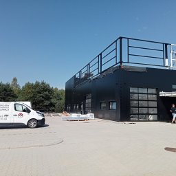 MAL-BAU WOŹNICA, KOWALCZYK SPÓŁKA CYWILNA - Czarny budynek po malowaniu elewacji, z widocznym autem firmy Mal-Gau i pracownikami. Elementy konstrukcji stalowej. Jasne, słoneczne ujęcie.
