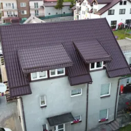 Dom z brązowym dachem z dachówki, widziany z góry, otoczony zielenią i zabudową. Szary budynek z oknami i kwiatami w skrzynkach. Ulica z boku, wieża z cegły.