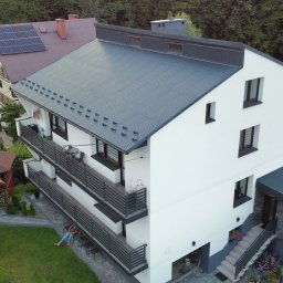 MAL-BAU WOŹNICA, KOWALCZYK SPÓŁKA CYWILNA - Nowoczesny, trzypiętrowy dom z szarą blachodachówką i białymi ścianami, widziany z góry. Na balkonie siedzi osoba. W tle zieleń drzew i drugi dom z panelami słonecznymi.