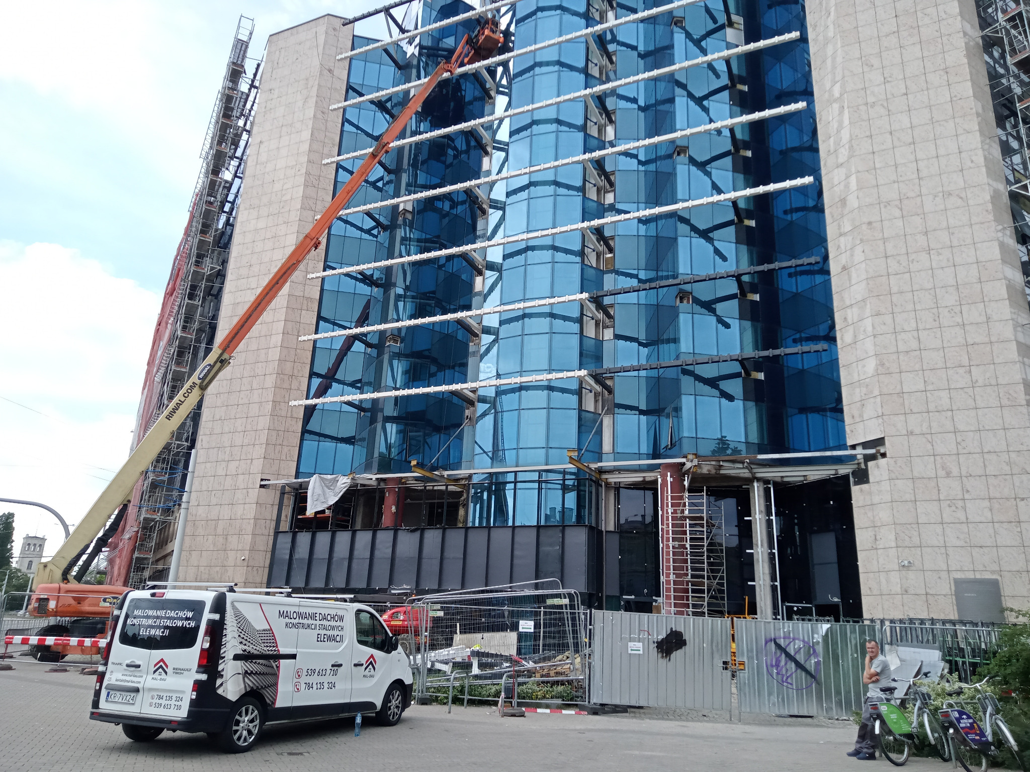 Prace na wysokościach przy szklanej elewacji biurowca. Podnośnik koszowy i rusztowania. Na pierwszym planie van z reklamą usług: malowanie dachów, elewacji i konstrukcji stalowych.