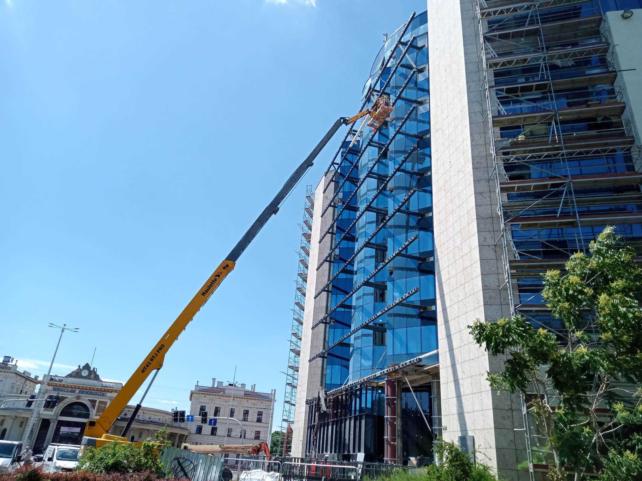 Praca na wysokościach: Żółty podnośnik koszowy przy szklanej elewacji biurowca w słoneczny dzień. Widoczne rusztowania i budynek dworca w tle.