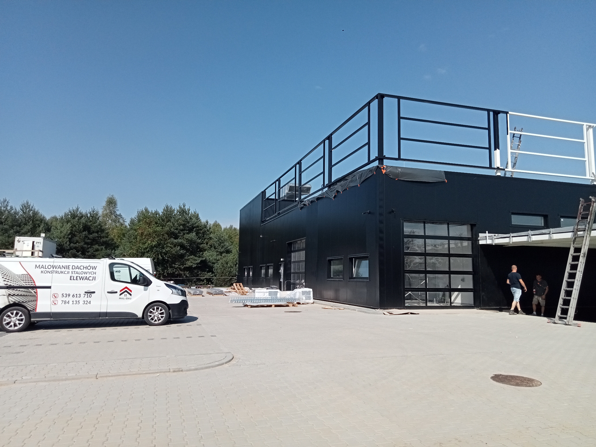 Czarny budynek po malowaniu elewacji, z widocznym autem firmy Mal-Gau i pracownikami. Elementy konstrukcji stalowej. Jasne, słoneczne ujęcie.
