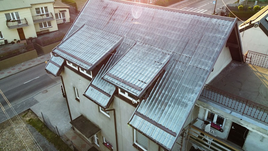 Zdjęcie z góry przedstawia dom z szarą dachówką i oknami. Dach jest częściowo pokryty mchem i wymaga czyszczenia. Widoczna ulica i inne budynki w tle.
