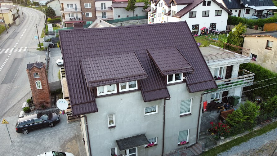 Dom z brązowym dachem z dachówki, widziany z góry, otoczony zielenią i zabudową. Szary budynek z oknami i kwiatami w skrzynkach. Ulica z boku, wieża z cegły.