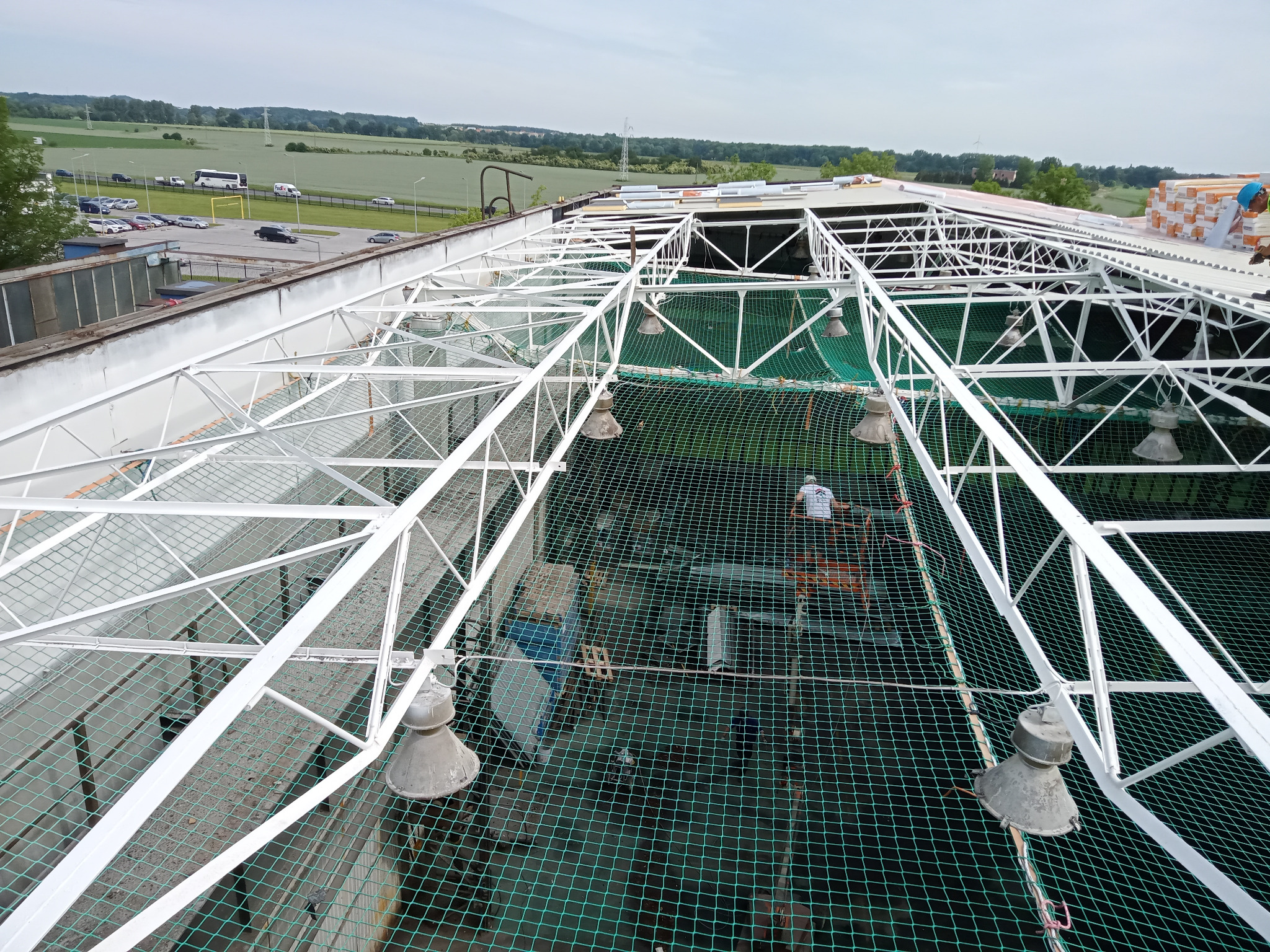 Widok z góry na konstrukcję dachu z siatką zabezpieczającą, pracownik w oddali. W tle budynki, parking i zielone pola. Jasne niebo.