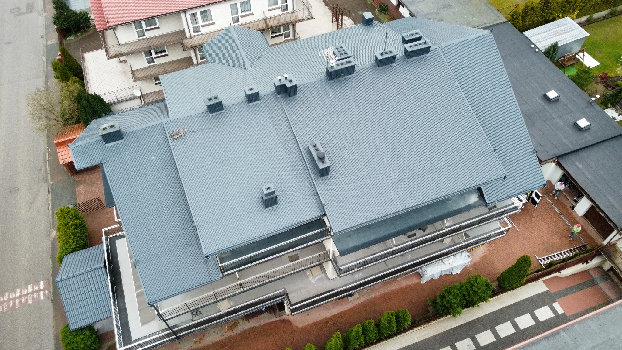 Metalowy dach wielospadowy po malowaniu w Witanowicach, widok z góry. Widoczne kominy i wyjścia wentylacyjne. Dach w kolorze szarym, otoczenie zielone.