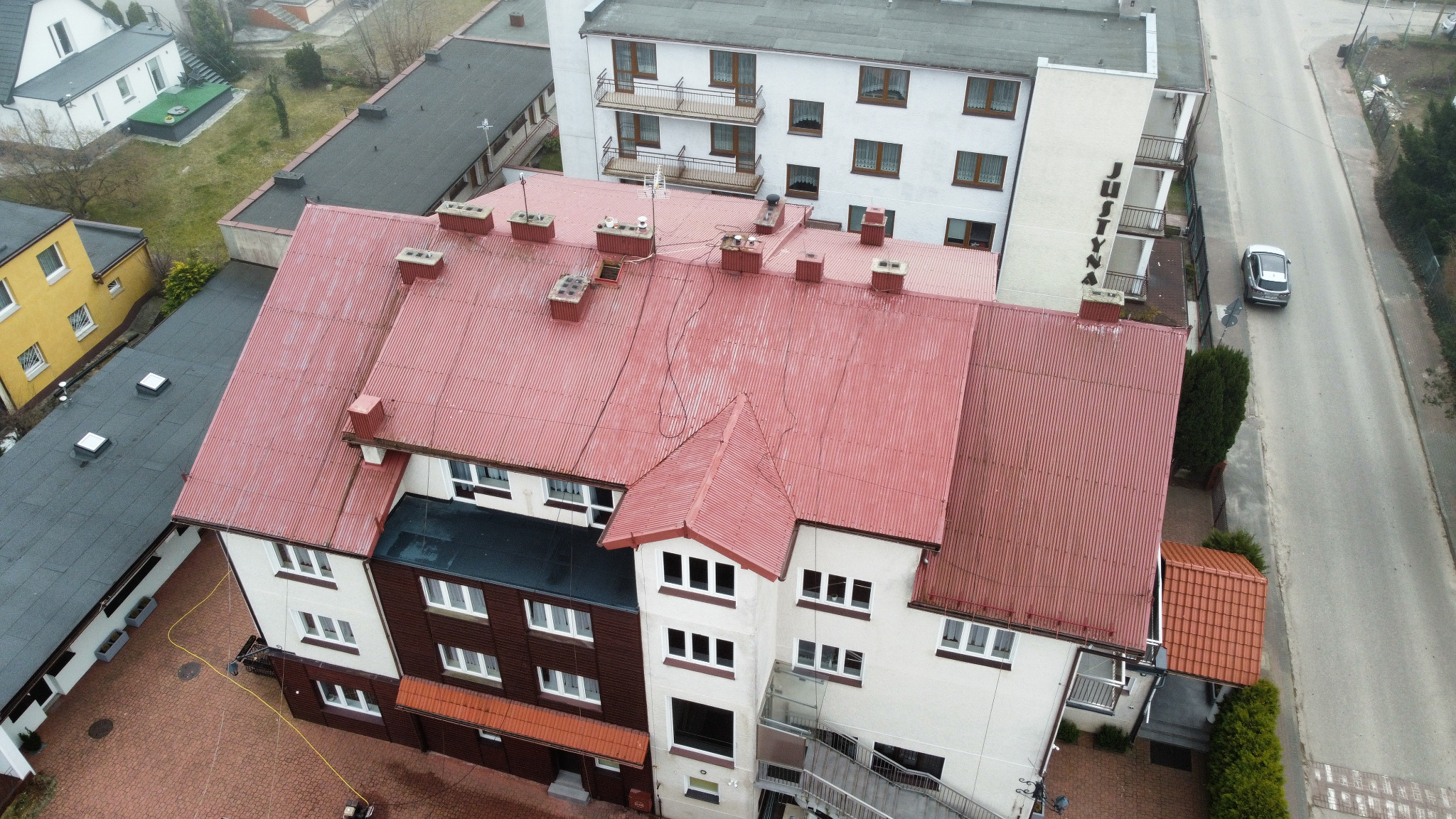 Czerwony, blaszany dach budynku wielorodzinnego z kominami, widziany z góry pod kątem. W tle zabudowa miejska i ulica z zaparkowanym samochodem.