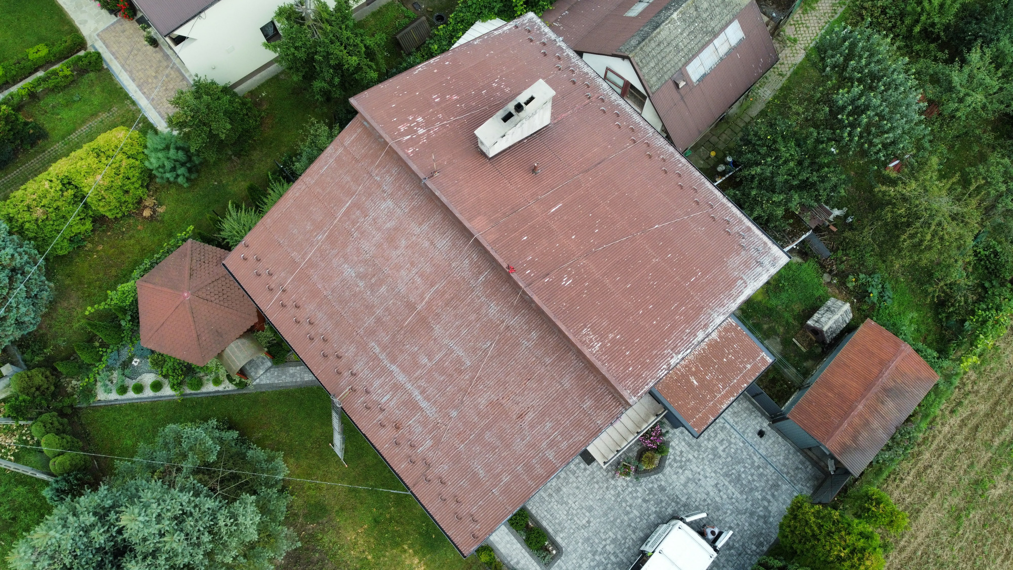 Widok z góry na dom z brązowym, lekko zabrudzonym dachem z kominem. Obok altana, kostka brukowa i zieleń. Częściowo widoczny samochód dostawczy.