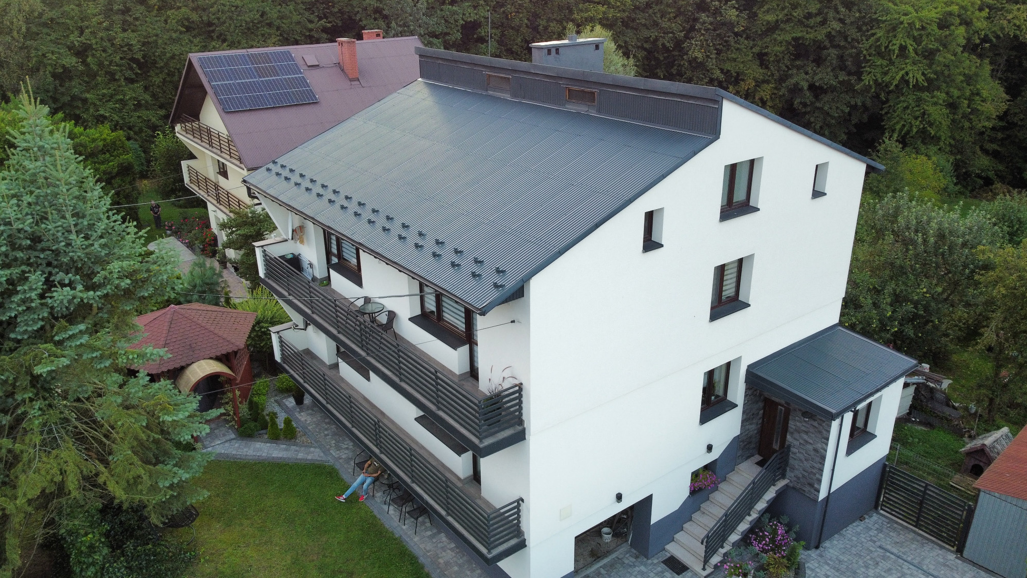 Nowoczesny, trzypiętrowy dom z szarą blachodachówką i białymi ścianami, widziany z góry. Na balkonie siedzi osoba. W tle zieleń drzew i drugi dom z panelami słonecznymi.