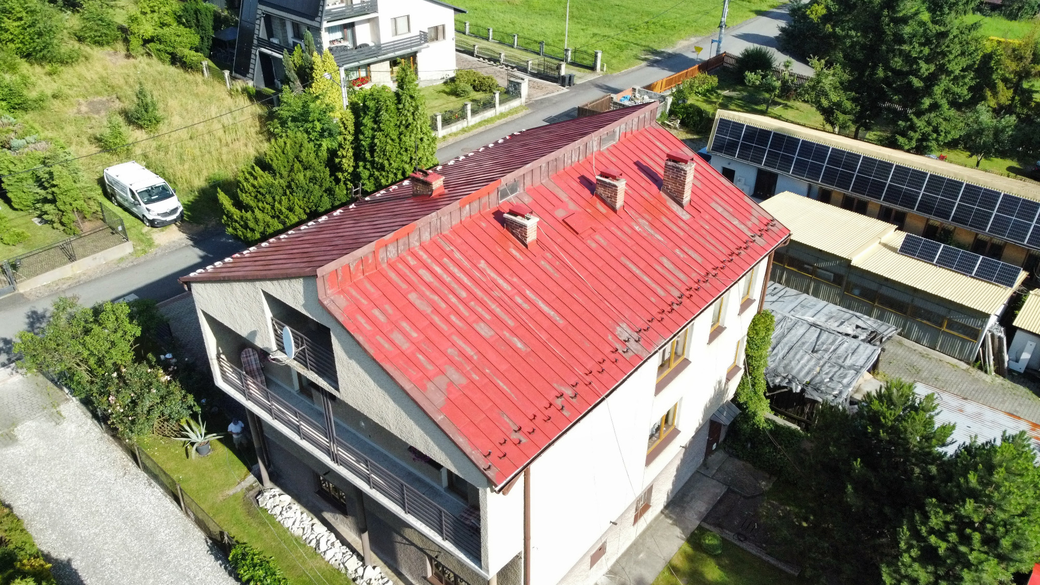Czerwony dach z blachy falistej na dwupiętrowym domu z balkonem, widziany z góry. Widoczne kominy, okna i otoczenie budynku z zielenią i zaparkowanym białym vanem.
