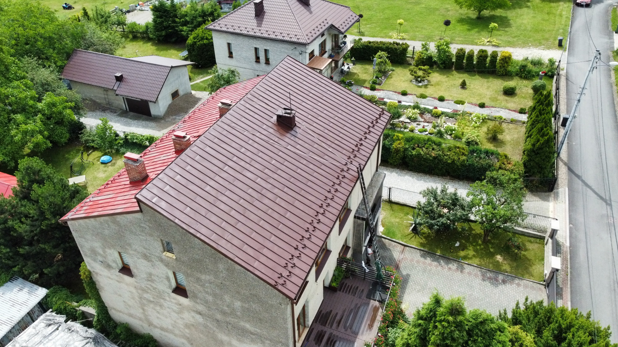 Widok z góry na dom z czerwonym dachem po renowacji. Widoczna drabina oparta o budynek, zadbany ogród i otoczenie. Czyste, nowe pokrycie dachowe.