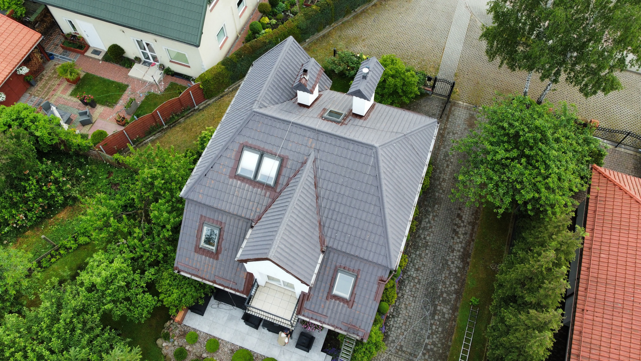 Widok z góry na dom z ciemnoszarym dachem, otoczony zielenią. Widoczne okna dachowe, kominy i fragmenty elewacji. Obok domu kostka brukowa i drzewa.