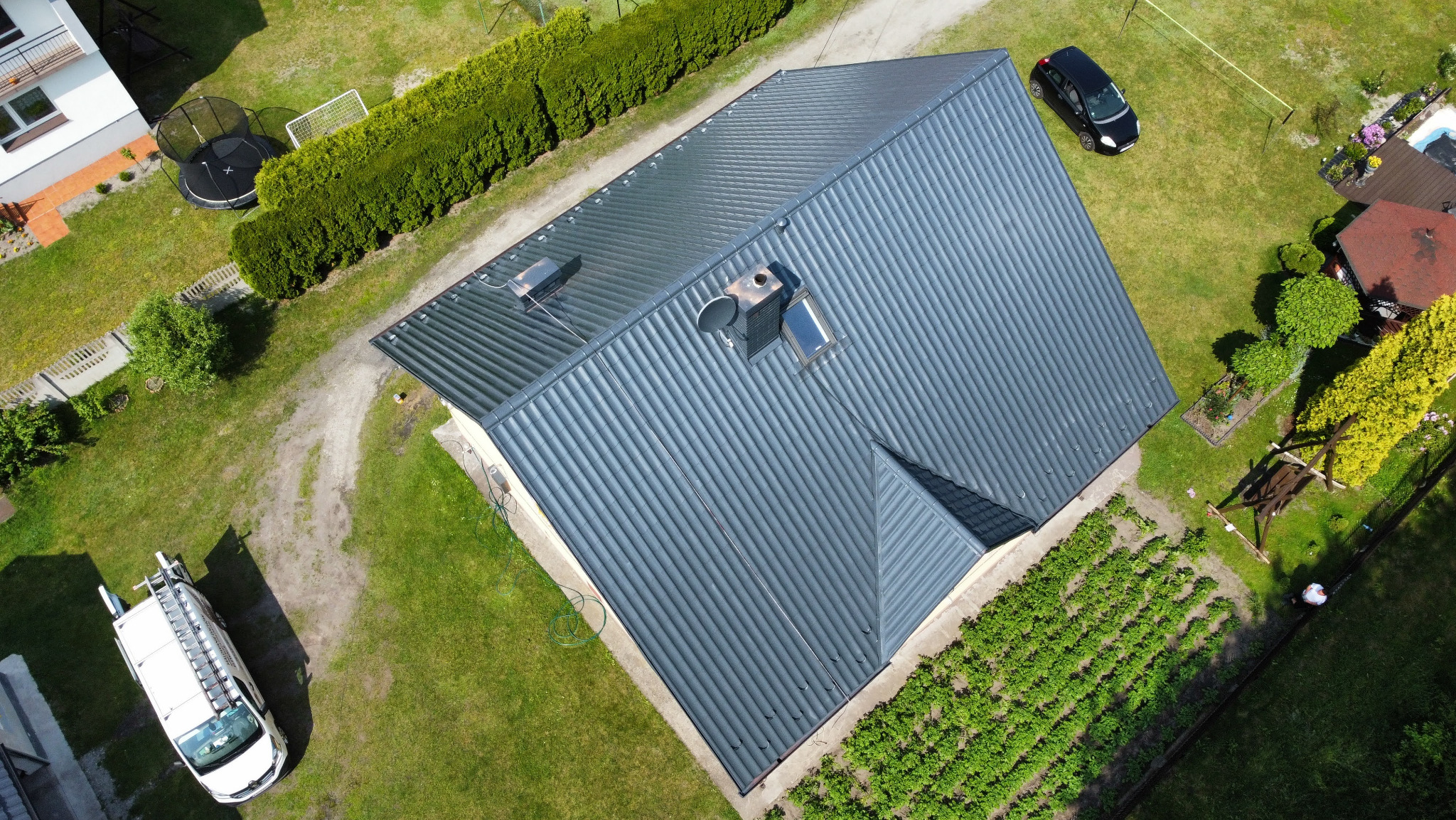 Zdjęcie z lotu ptaka przedstawia dom z ciemnoszarym, odnowionym dachem z blachy trapezowej, z kominem i oknami dachowymi. Obok zaparkowany samochód i bus z drabiną.