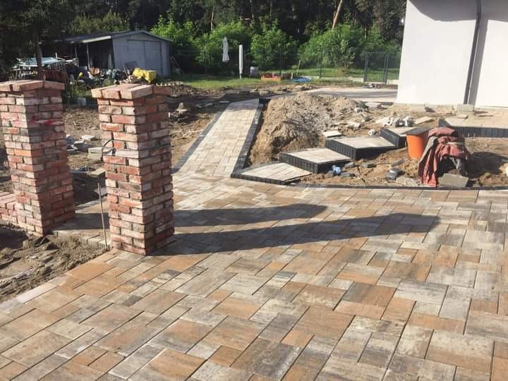 Układanie kostki brukowej: wzór jodełki, dwa ceglane słupki na wejściu, schody z kostki prowadzące do domu, widoczne narzędzia i materiały budowlane.
