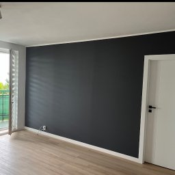 Sebastian Tramś Usługi remontowo-wykończeniowe - Minimalistyczne wnętrze: ciemna ściana kontrastuje z jasną podłogą i drzwiami. Widok na balkon z roślinnością. Nowoczesne oświetlenie punktowe.