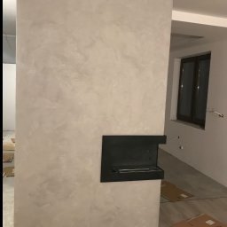 Sebastian Tramś Usługi remontowo-wykończeniowe - Ściana z dekoracyjną, szarą strukturą i wbudowanym biokominkiem, wykończona białą listwą przypodłogową. Widoczne kartony na podłodze i fragment okna.