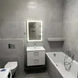 Nowoczesna łazienka w szarościach: umywalka z szafką, podświetlane lustro, wanna i toaleta. Minimalistyczny design, płytki imitujące beton, pralka częściowo widoczna w kadrze.