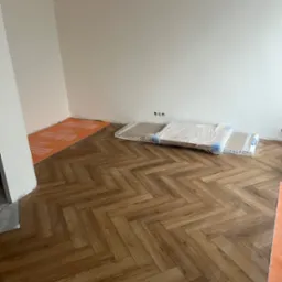 Ukończona podłoga z paneli ułożonych w jodełkę, widoczne materiały przygotowane do dalszych prac wykończeniowych, pomarańczowa mata na podłodze.