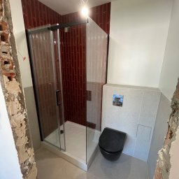 Sebastian Tramś Usługi remontowo-wykończeniowe - Nowoczesna łazienka w trakcie wykańczania: kabina prysznicowa z czarną ramą, wisząca toaleta i bordowe płytki na ścianie. Widoczne surowe fragmenty ścian.