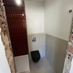 Sebastian Tramś Usługi remontowo-wykończeniowe - Wnętrze łazienki w trakcie prac wykończeniowych. Widoczna kabina prysznicowa z bordowymi płytkami, podwieszana toaleta i częściowo położone płytki na ścianach.