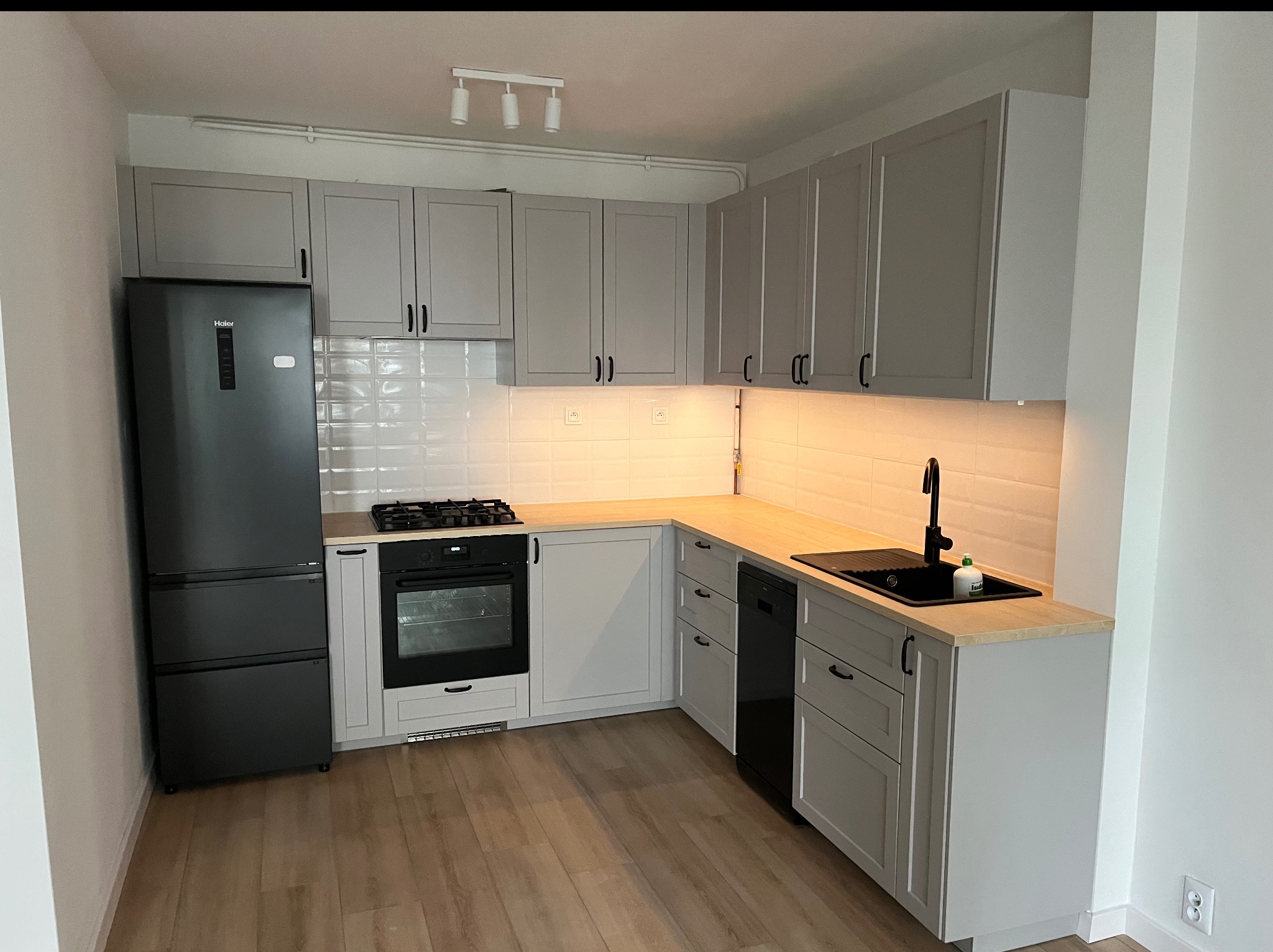 Jasnoszara kuchnia po remoncie: zabudowa z czarnymi uchwytami, drewniany blat, białe płytki na ścianie, czarny zlew i sprzęt AGD. Nowoczesny design.