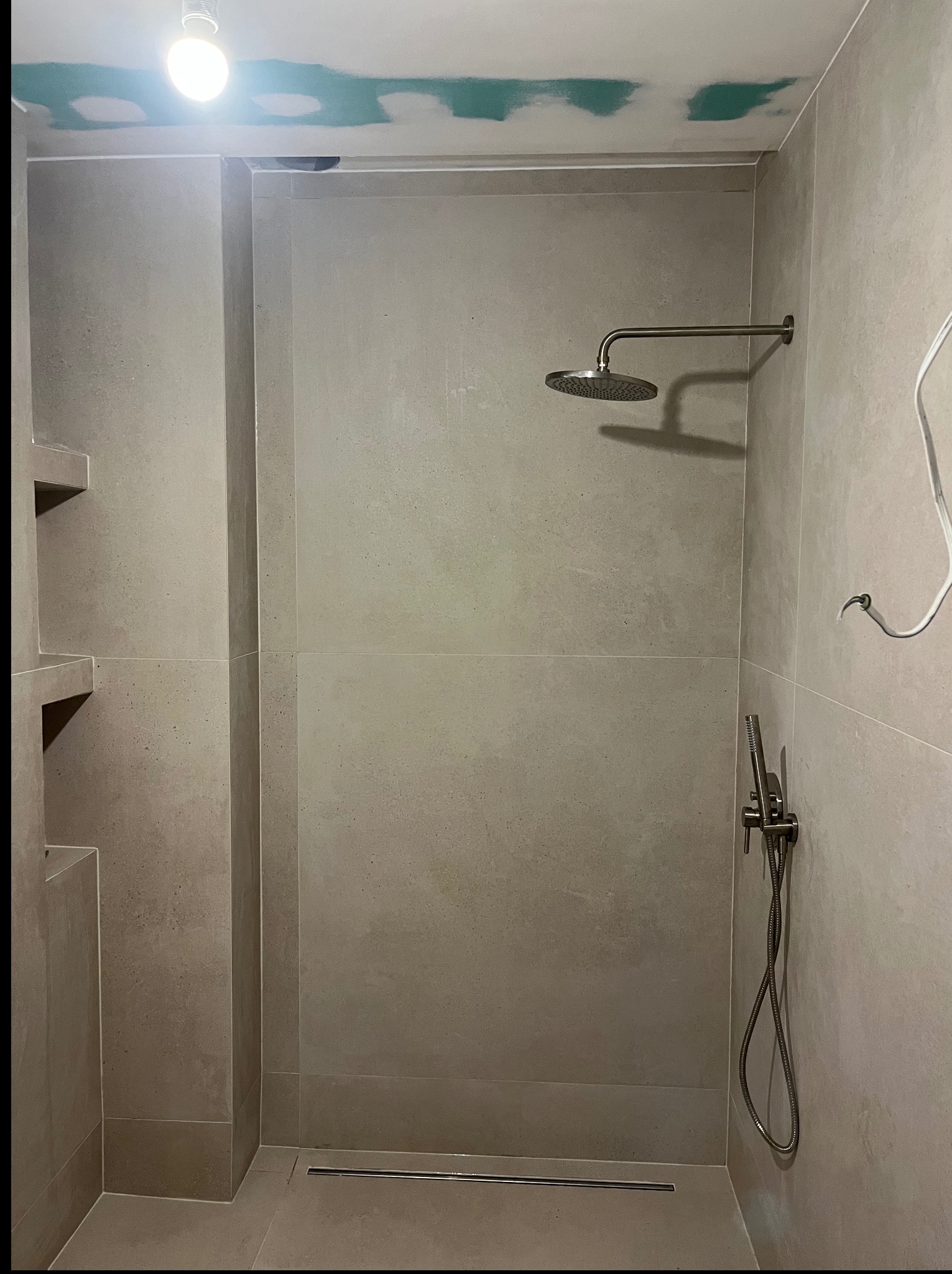 Nowoczesna kabina prysznicowa z szarymi płytkami, deszczownicą i odpływem liniowym. Sufit z widocznymi śladami po szpachlowaniu. Minimalistyczny design.