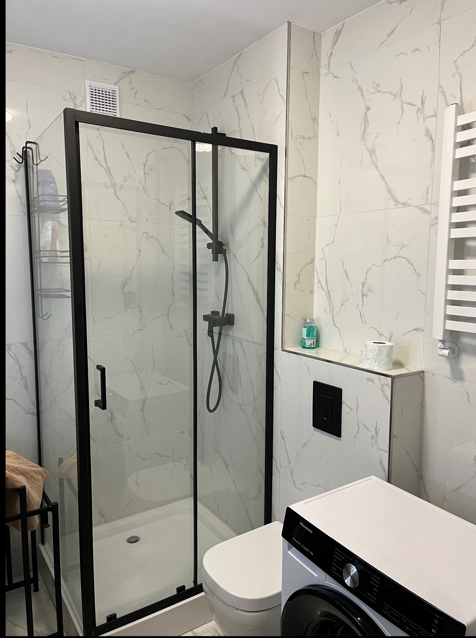 Wykończona łazienka z kabiną prysznicową typu walk-in z czarnymi profilami, białe płytki imitujące marmur, wisząca toaleta i pralka. Nowoczesny design.