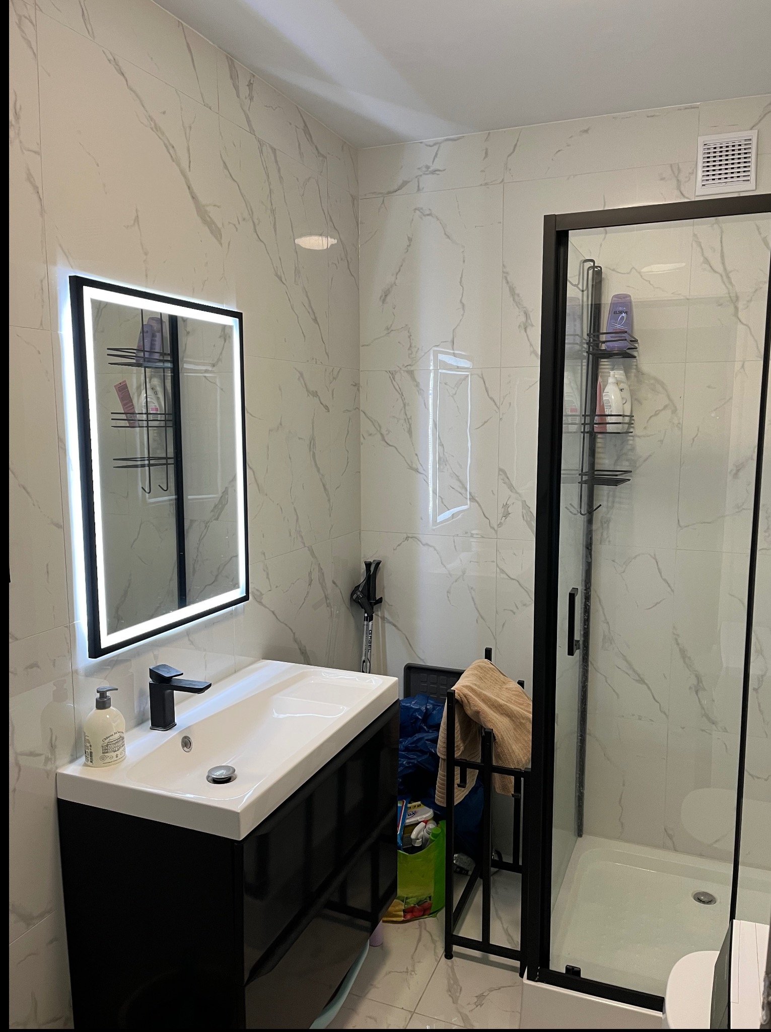 Nowoczesna łazienka z kabiną prysznicową typu walk-in i podświetlanym lustrem LED. Białe płytki imitujące marmur na ścianach i czarne akcenty armatury.