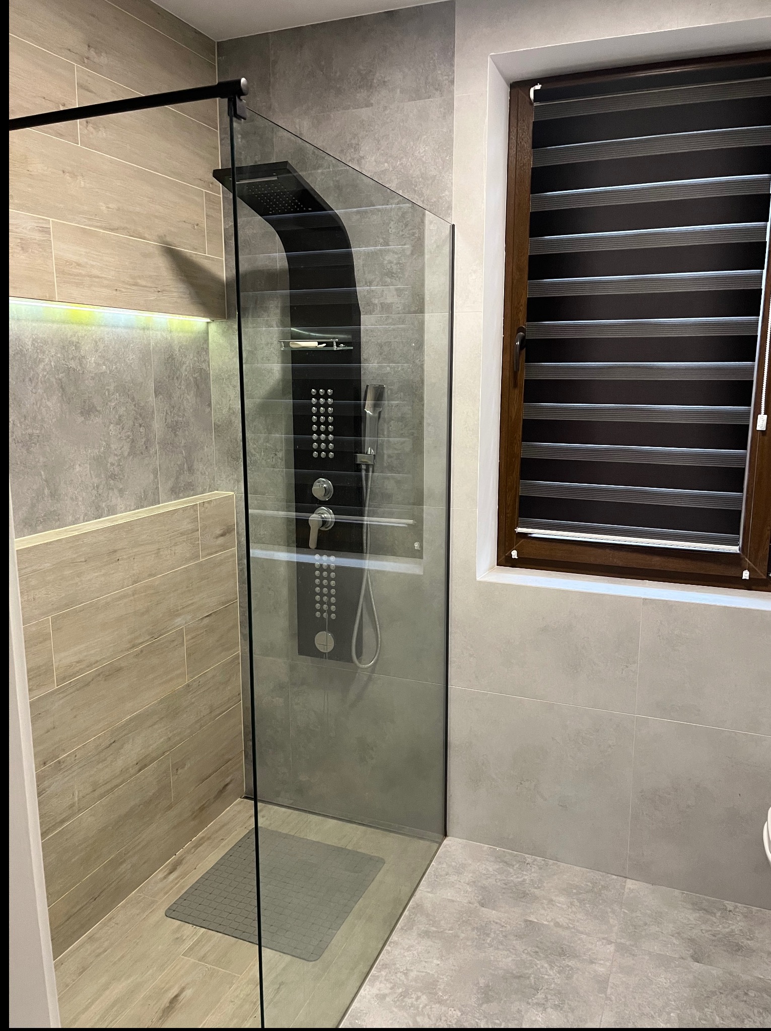 Nowoczesna kabina prysznicowa typu walk-in z czarnym panelem natryskowym i szklaną ścianką, wykończona płytkami w odcieniach szarości i beżu, z oknem z żaluzjami.