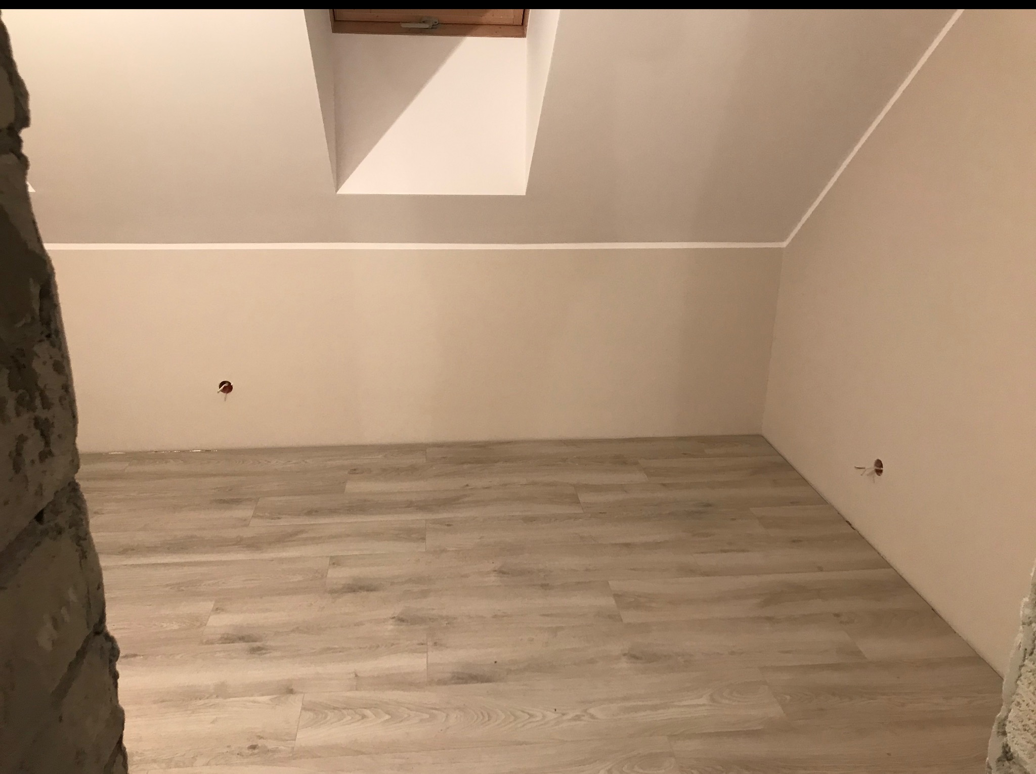 Poddasze po remoncie: jasne panele podłogowe, świeżo pomalowane ściany w odcieniach beżu, okno dachowe, widoczne puszki elektryczne. Częściowo widoczna ściana z cegły.