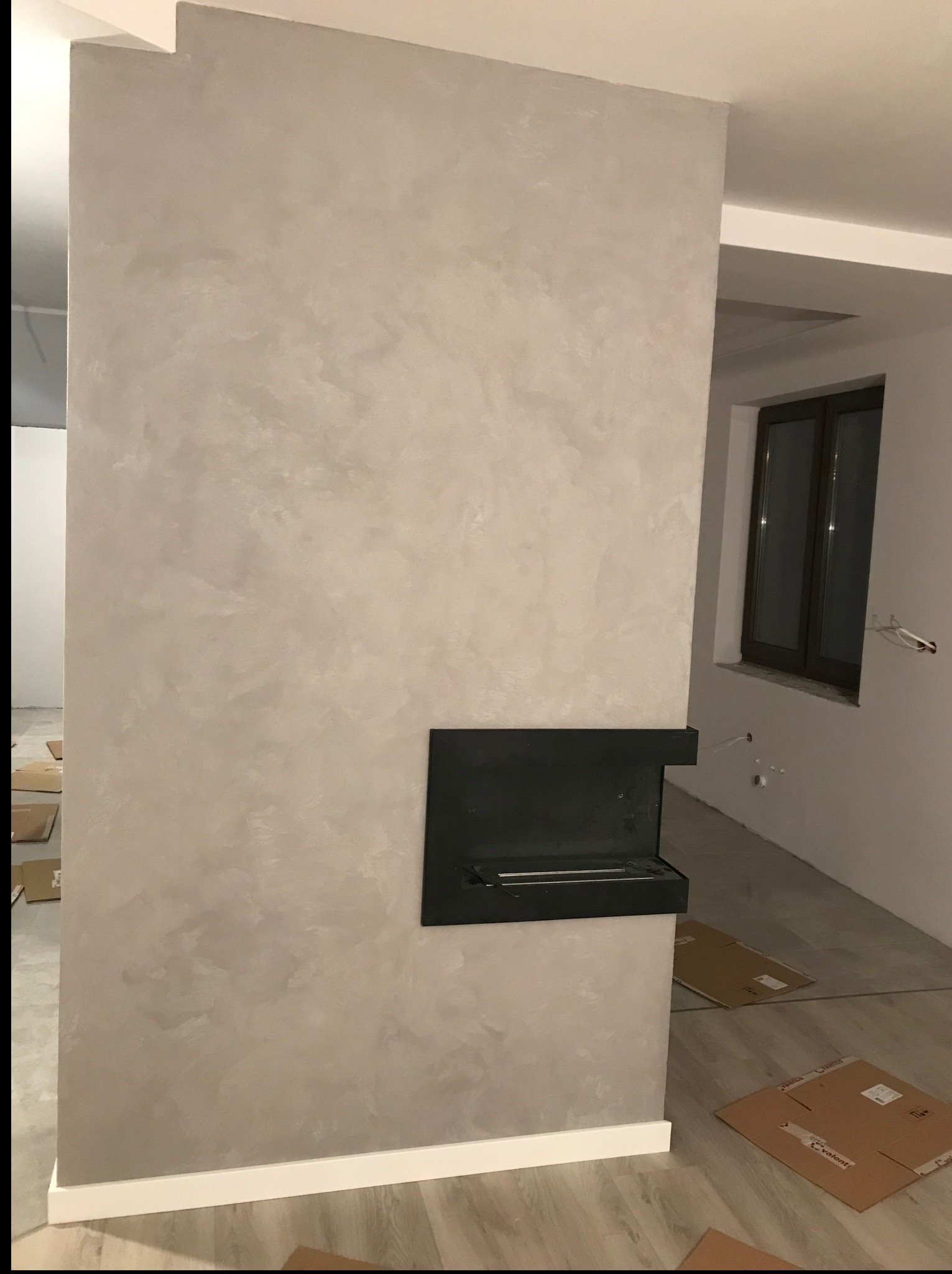 Ściana z dekoracyjną, szarą strukturą i wbudowanym biokominkiem, wykończona białą listwą przypodłogową. Widoczne kartony na podłodze i fragment okna.