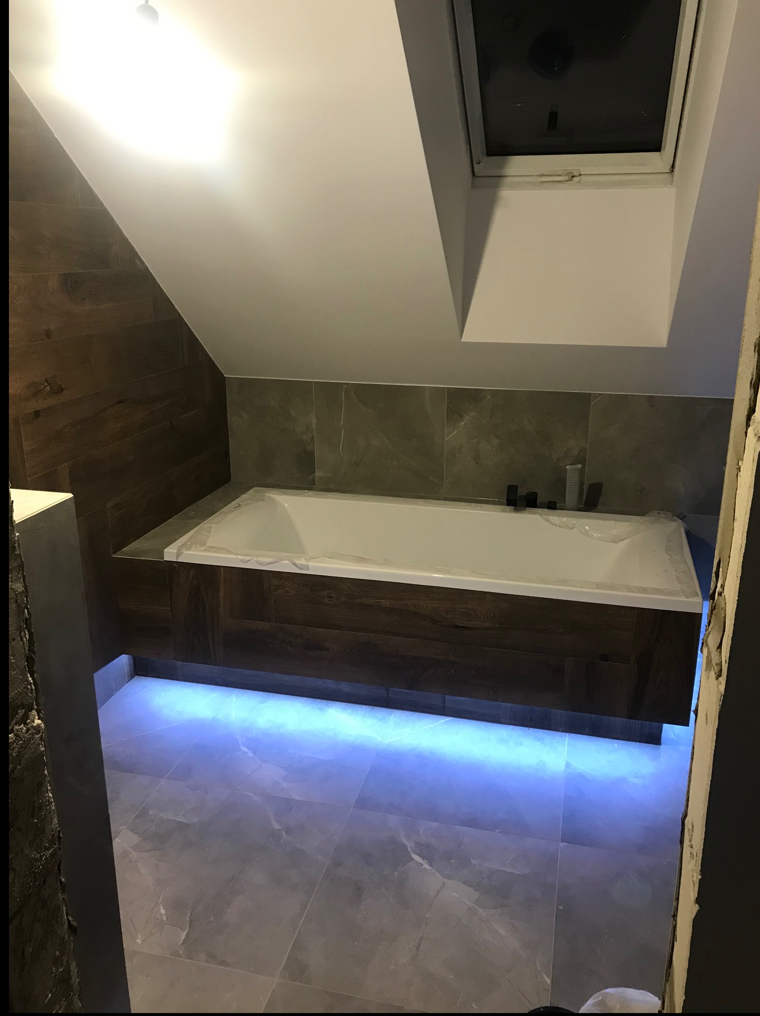 Nowoczesna łazienka na poddaszu z oknem dachowym i podświetlaną wanną obudowaną drewnem. Ułożone płytki podłogowe i ścienne imitujące drewno i kamień.