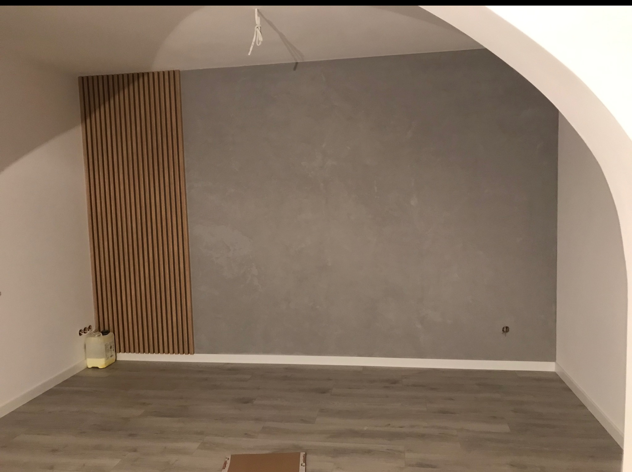 Minimalistyczne wnętrze po remoncie: szara ściana z dekoracyjną strukturą, drewniane lamele na ścianie i jasna podłoga. Widoczny fragment łuku architektonicznego.