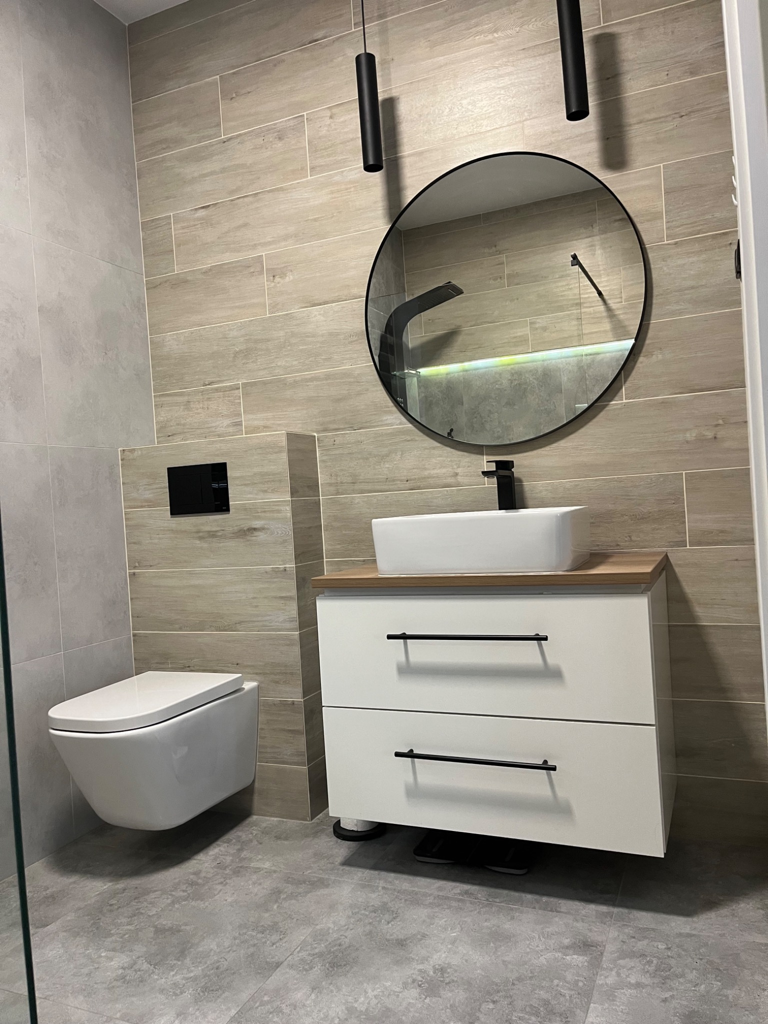 Nowoczesna łazienka w Prażmowie z podwieszaną toaletą i umywalką nablatową. Płytki imitujące drewno na ścianach i szare na podłodze. Czarne akcenty.