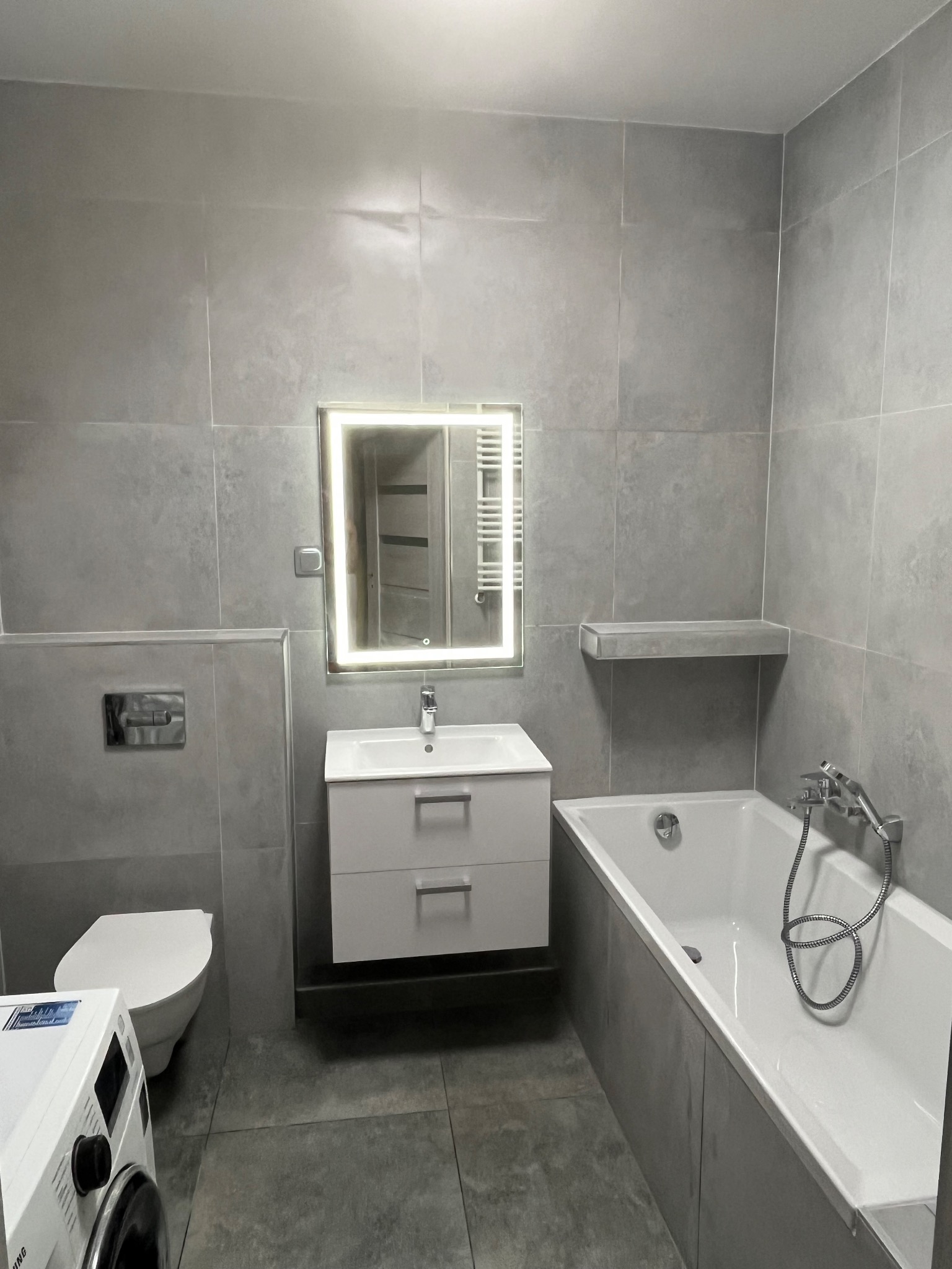 Nowoczesna łazienka w szarościach: umywalka z szafką, podświetlane lustro, wanna i toaleta. Minimalistyczny design, płytki imitujące beton, pralka częściowo widoczna w kadrze.
