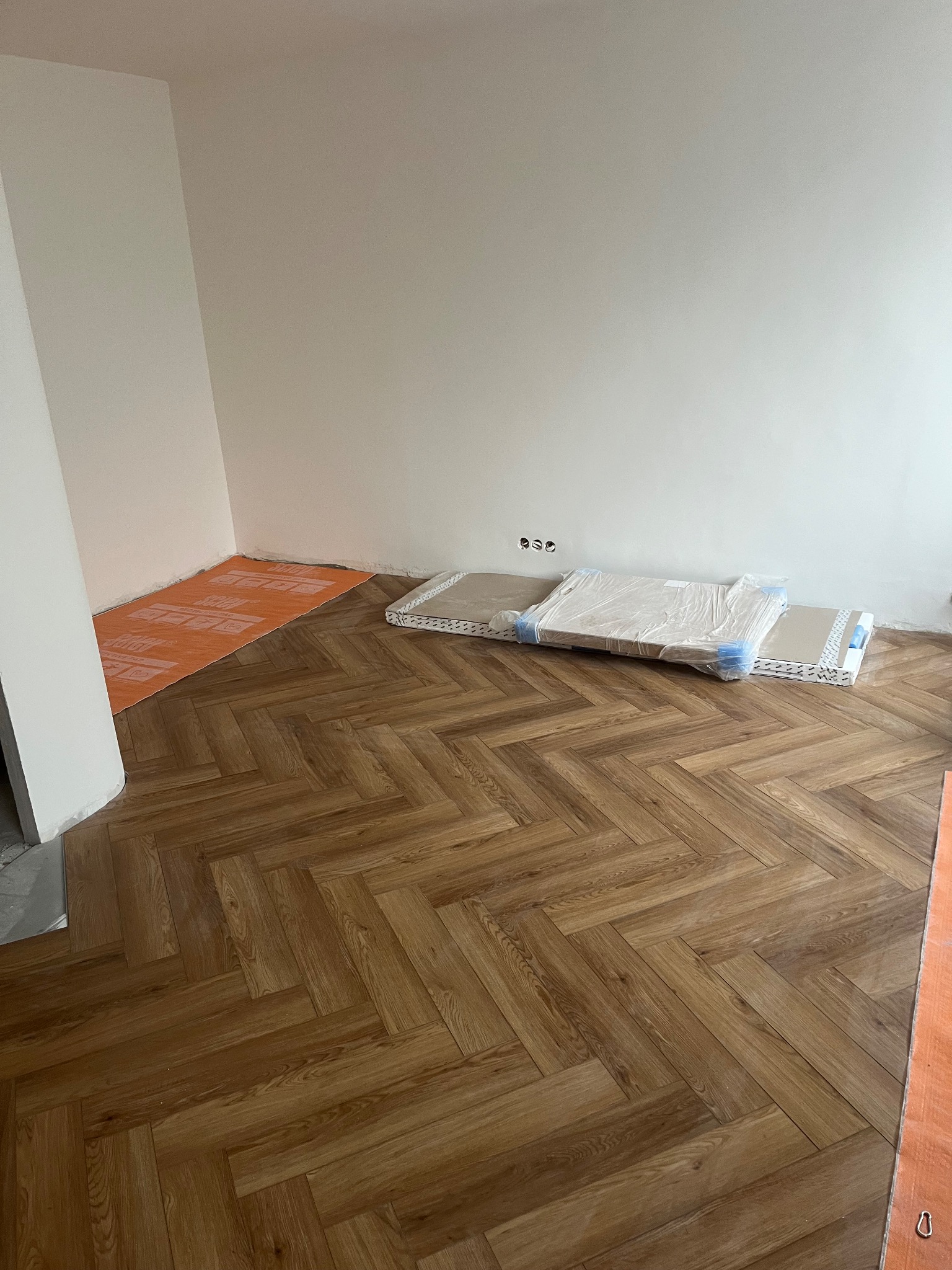 Ukończona podłoga z paneli ułożonych w jodełkę, widoczne materiały przygotowane do dalszych prac wykończeniowych, pomarańczowa mata na podłodze.