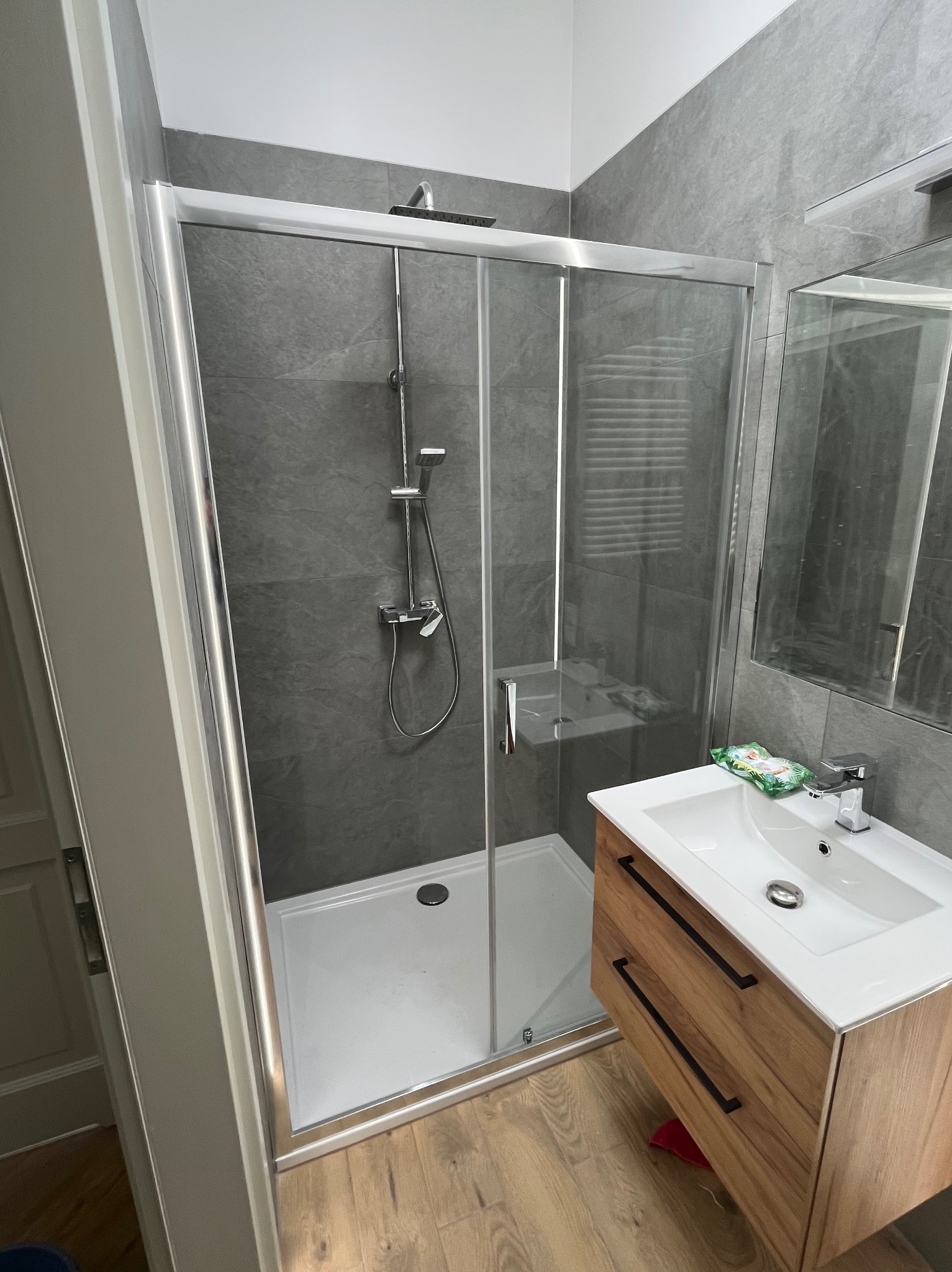 Nowoczesna łazienka z kabiną prysznicową typu walk-in, szare płytki imitujące kamień i drewniana szafka pod umywalką z czarnymi uchwytami. Minimalistyczny design.